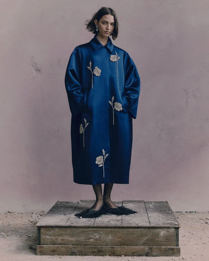 Model standing on a wooden pedestal wearing a long blue coat with embroidered floral details against a muted backdrop.