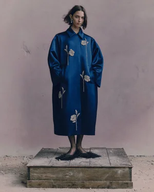 Model standing on a wooden pedestal wearing a long blue coat with embroidered floral details against a muted backdrop.