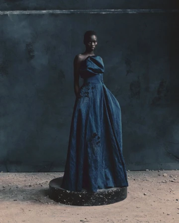 Model standing on a circular plinth wearing a dark structured gown against a mottled dark wall