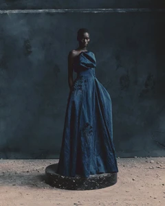 Model standing on a circular plinth wearing a dark structured gown against a mottled dark wall