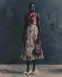 Model wearing a floral print sleeveless top and pleated skirt with long burgundy leather gloves standing against a dark wall