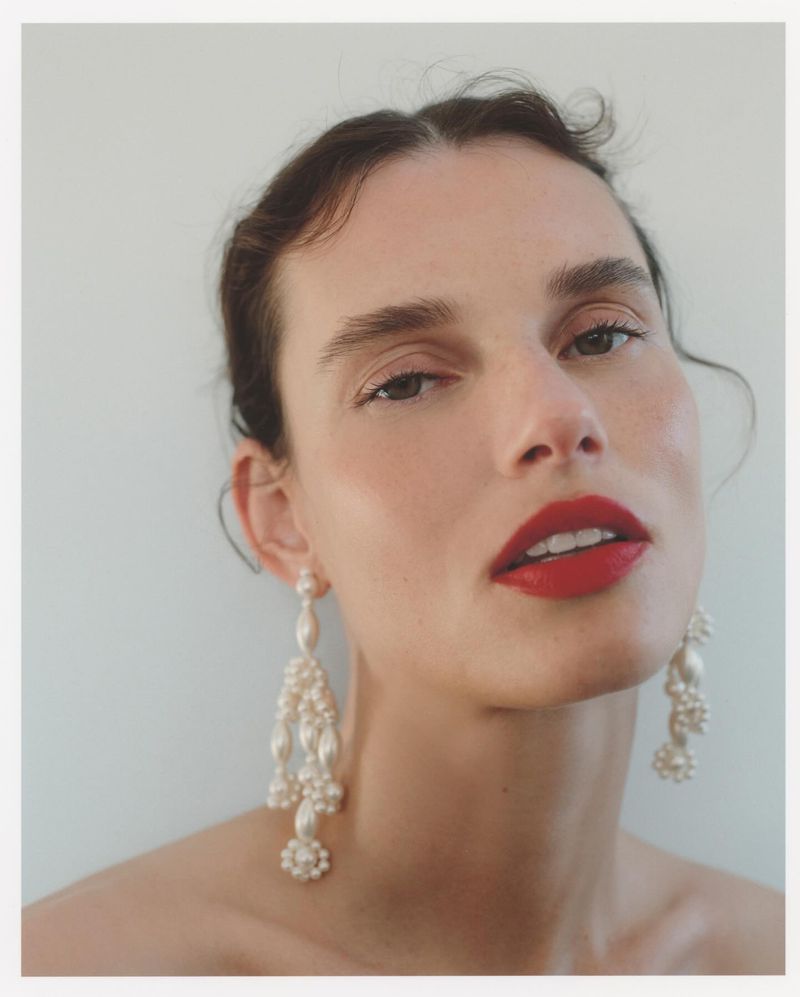 Close-up portrait of a fair-skinned woman with red lipstick and pearl earrings against a light background in a beauty shot.