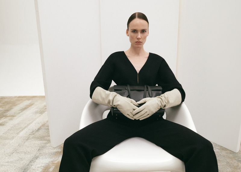 Elegant woman sitting in a chair, holding a black leather handbag, wearing long gloves, and a black jumpsuit in a modern white room.