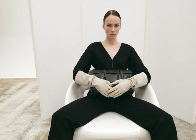 Elegant woman sitting in a chair, holding a black leather handbag, wearing long gloves, and a black jumpsuit in a modern white room.