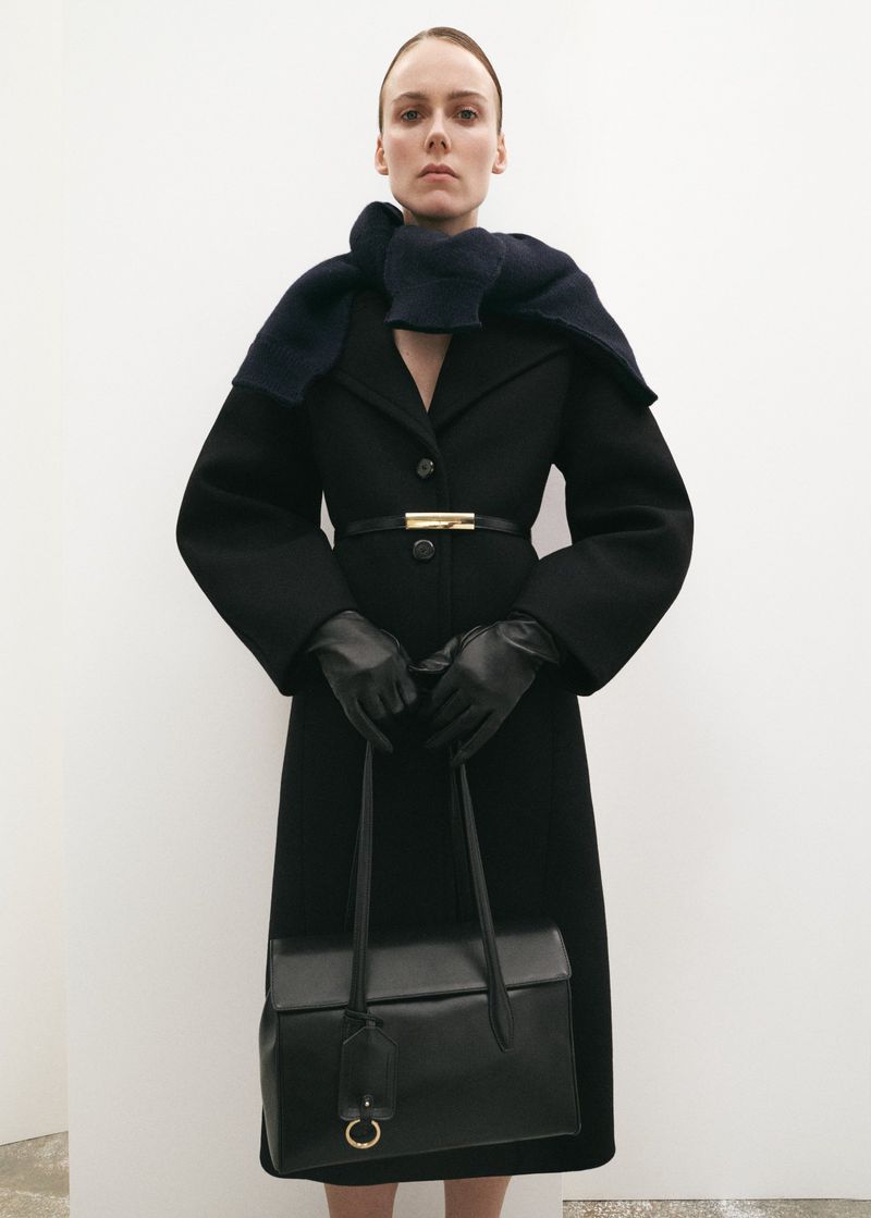 Elegant woman in black coat, scarf, and leather gloves holding a structured leather handbag against a plain white background.