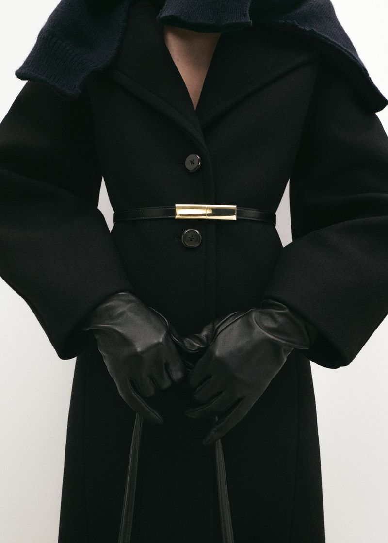 A person wearing a black coat with a thin black belt and leather gloves is captured in a studio shot, showcasing a fashionable winter outfit.