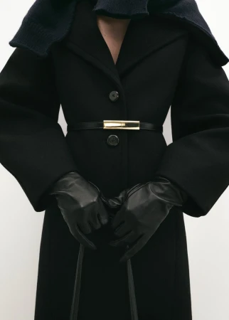 A person wearing a black coat with a thin black belt and leather gloves is captured in a studio shot, showcasing a fashionable winter outfit.