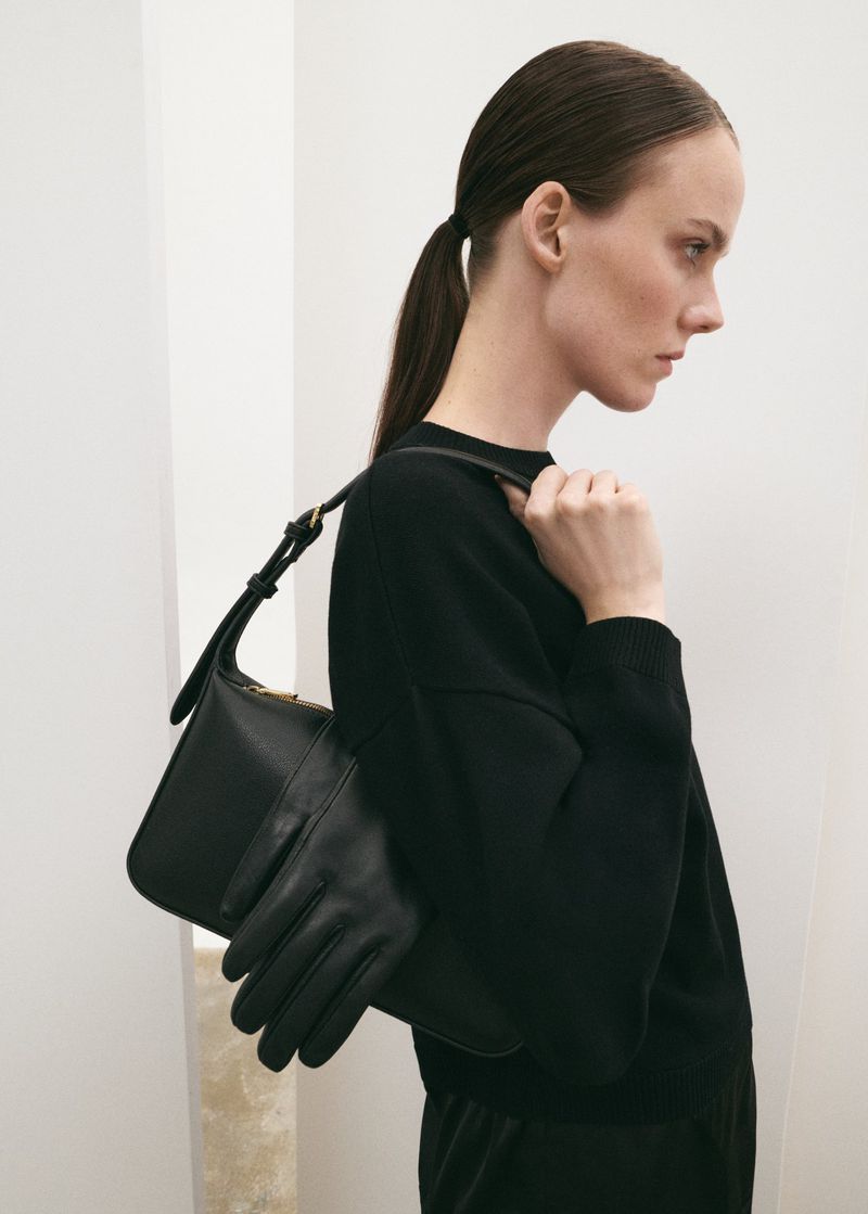 A woman in a black sweater and gloves carries a black leather handbag in a studio shot against a white backdrop. Fashion editorial.