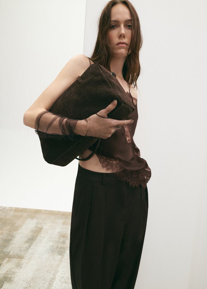 A woman with long brown hair holds a dark brown suede handbag and poses in a studio with a white backdrop. She wears a brown lace top.
