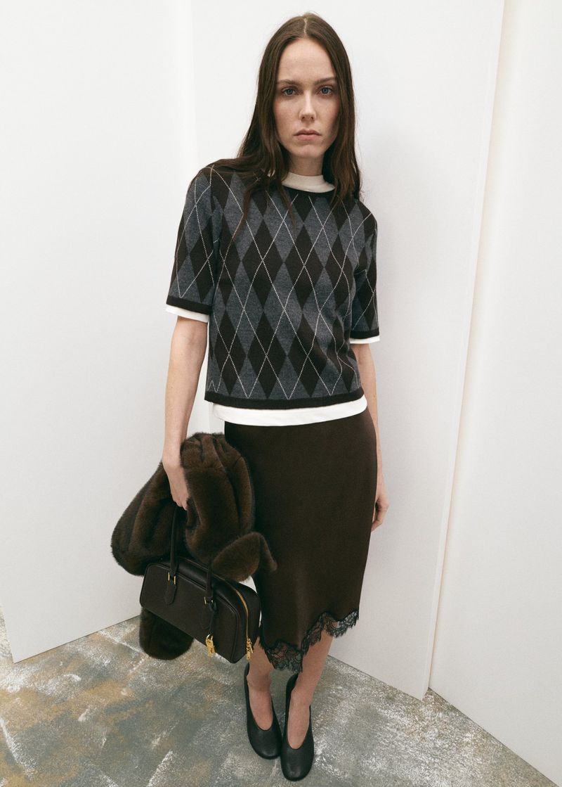 Studio shot of a female model in a patterned sweater and skirt, holding a handbag with a fur accessory, against a white wall.