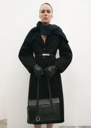 Model Kiki Willems wearing a black tailored coat with belted waist, black leather gloves, and holding a black leather tote bag.