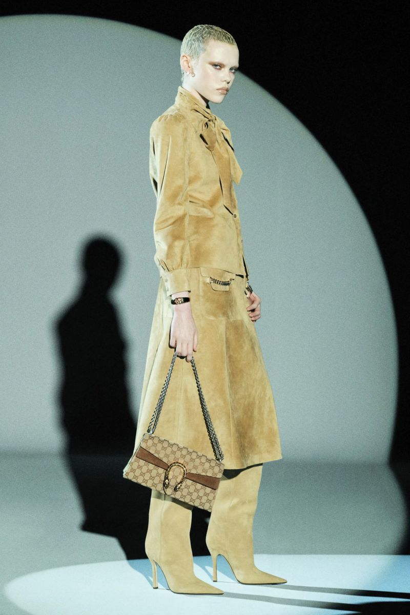 A person in a tan skirt suit and boots, accessorized with jewelry and a Gucci purse, is posing in a studio setting with a spotlight.