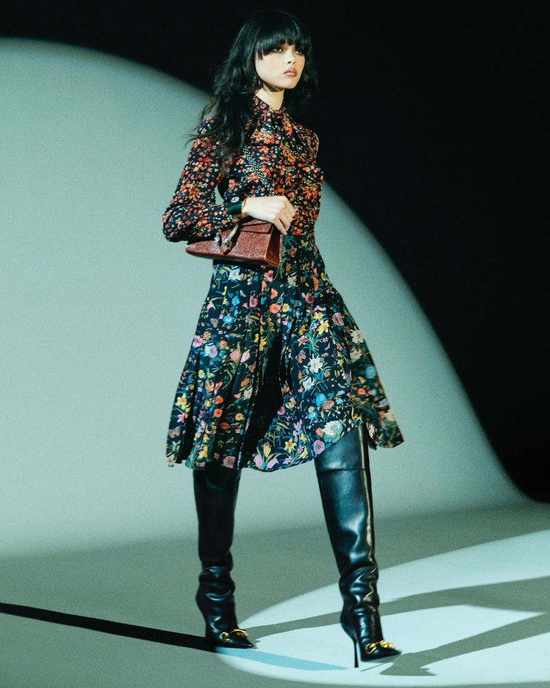 Model in a floral print dress and black leather boots holding a brown handbag under a spotlight in a studio.