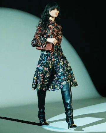 Model in a floral print dress and black leather boots holding a brown handbag under a spotlight in a studio.