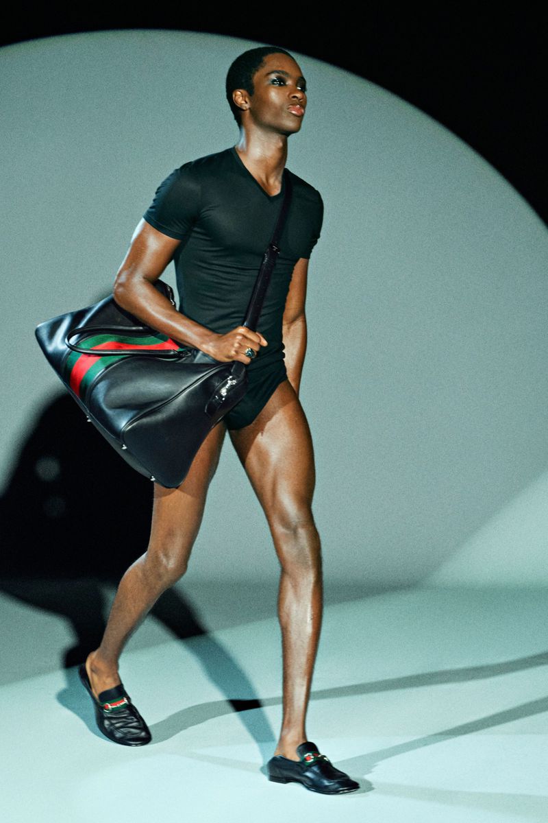 A Black male model showcases black athletic wear and a black duffel bag with red and green stripes on a runway in a fashion show.