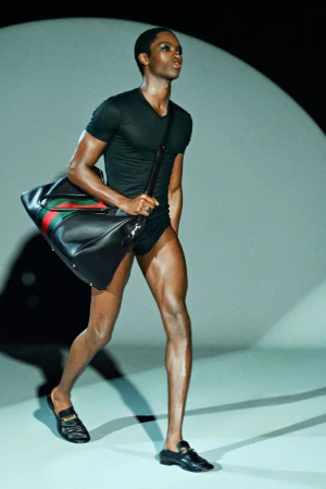 A Black male model showcases black athletic wear and a black duffel bag with red and green stripes on a runway in a fashion show.
