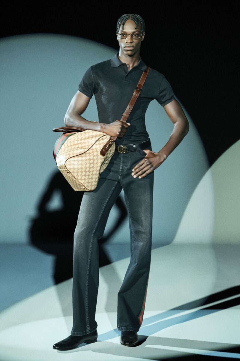 A male fashion model poses in dark jeans and a polo shirt, carrying a Gucci bag in a studio environment with soft, directional lighting.