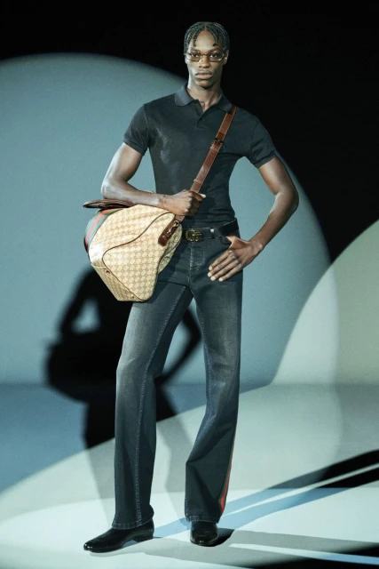 A male fashion model poses in dark jeans and a polo shirt, carrying a Gucci bag in a studio environment with soft, directional lighting.