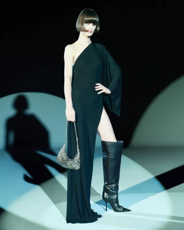 Model posing in a black one-shoulder high-slit gown and tall leather boots holding a monogram chain handbag under spotlight.