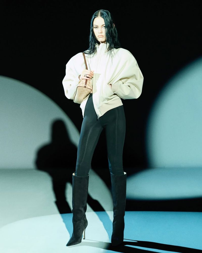 A model poses in a studio setting wearing a jacket, leggings, and boots, illuminated by spotlighting on a gradient backdrop.
