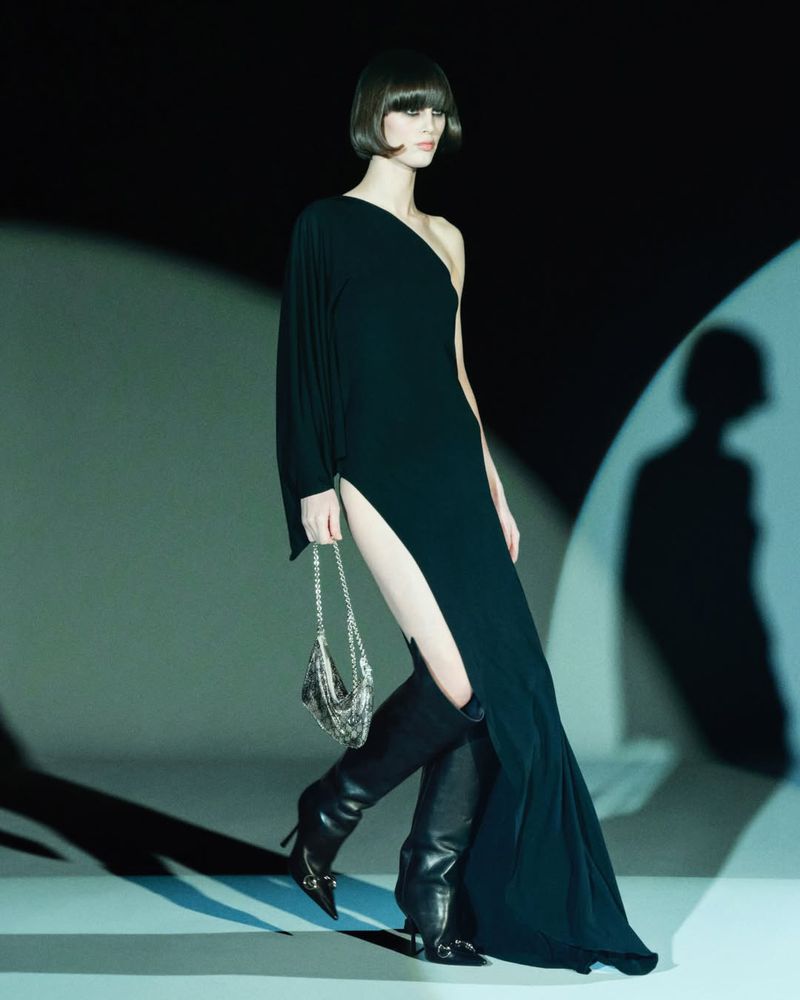 Elegant woman in a one-shoulder black gown and high boots strikes a pose against a subtly lit backdrop, holding a beaded purse.