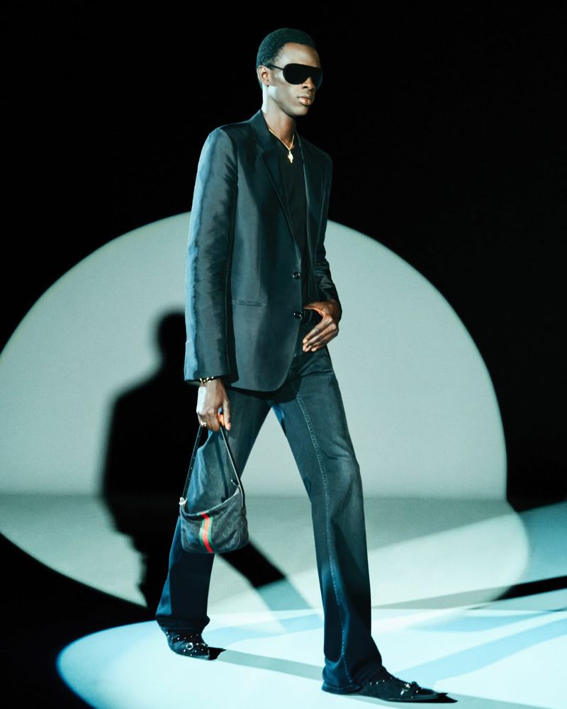A stylish black man wearing a dark suit, sunglasses, and carrying a handbag, walking under a spotlight on a fashion runway.