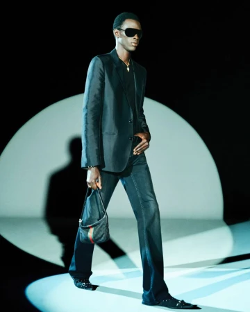 A stylish black man wearing a dark suit, sunglasses, and carrying a handbag, walking under a spotlight on a fashion runway.
