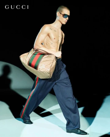 Male model walks on a runway with a Gucci duffel bag, wearing sunglasses and blue track pants with a green and red stripe.
