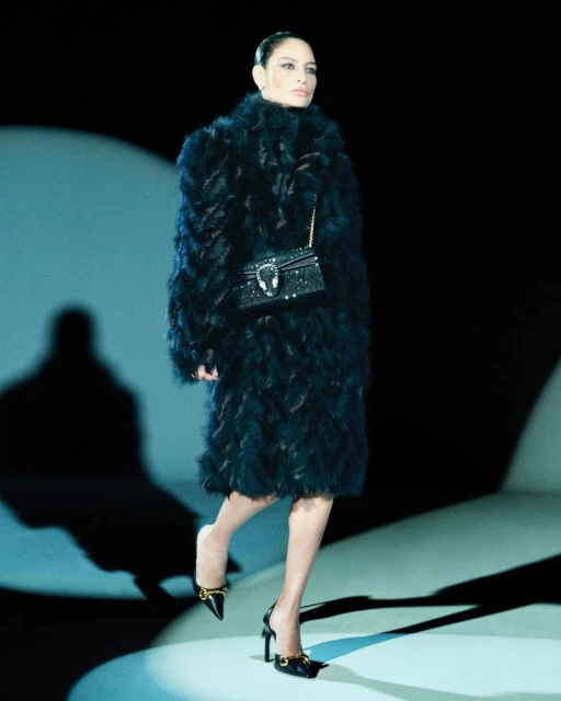 A woman in a dark feather coat walks the runway with a small, ornate shoulder bag and black high heel shoes on a softly lit runway.