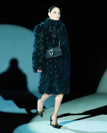 A woman in a dark feather coat walks the runway with a small, ornate shoulder bag and black high heel shoes on a softly lit runway.