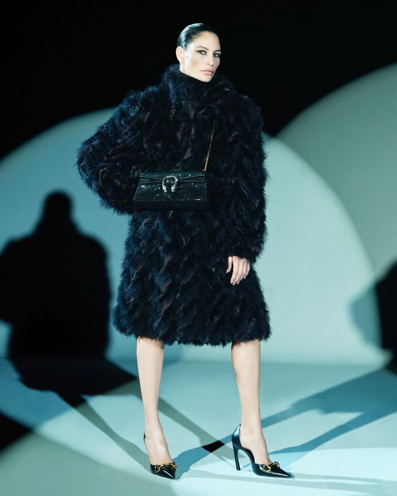 Model wearing a textured black feather coat, black chain shoulder bag, and black pointed-toe heels against a studio spotlight backdrop.