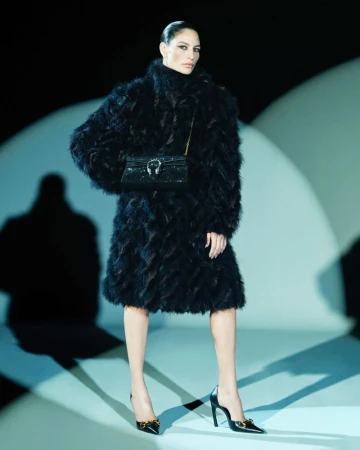 Model wearing a textured black feather coat, black chain shoulder bag, and black pointed-toe heels against a studio spotlight backdrop.