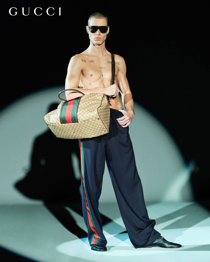 A shirtless male fashion model in sunglasses carries a Gucci travel bag on a runway, showcasing modern style against a dark background.