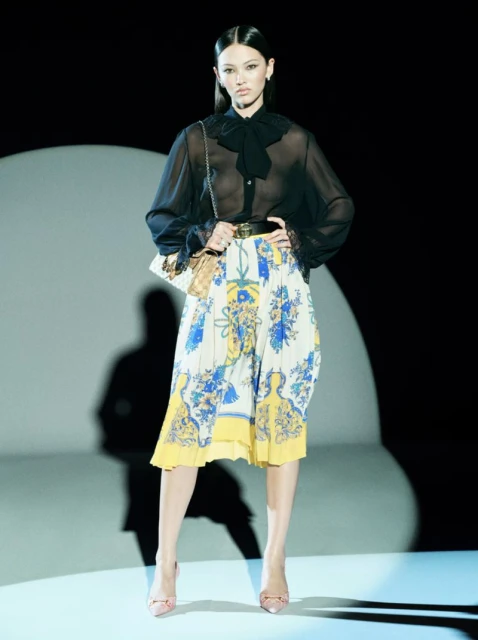 Elegant woman poses in a sheer black blouse, floral skirt, and heels, accented by a handbag under a spotlight against a dark background.