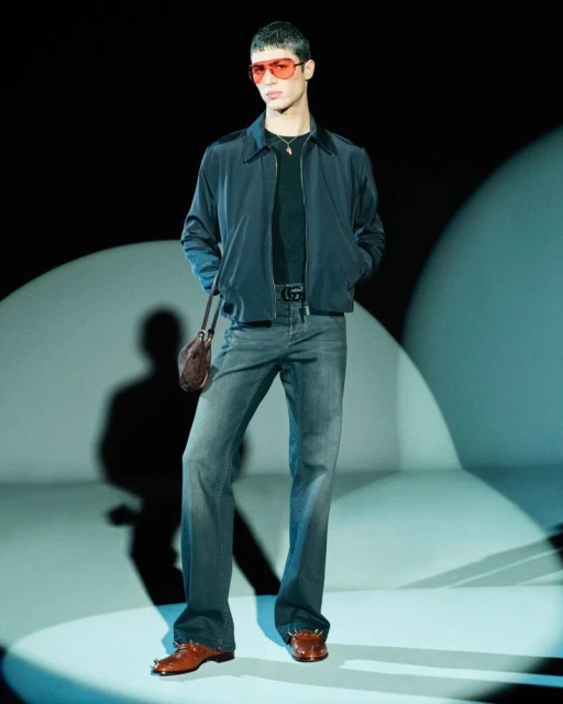 Full-length studio shot of a young adult male model in a dark jacket, jeans, and brown dress shoes, wearing red sunglasses, hands in his pockets.
