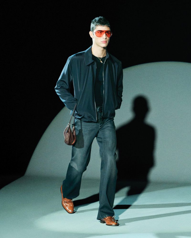 A male model walks on a runway wearing a blue jacket, grey jeans, brown shoes, and red sunglasses in a fashion show.