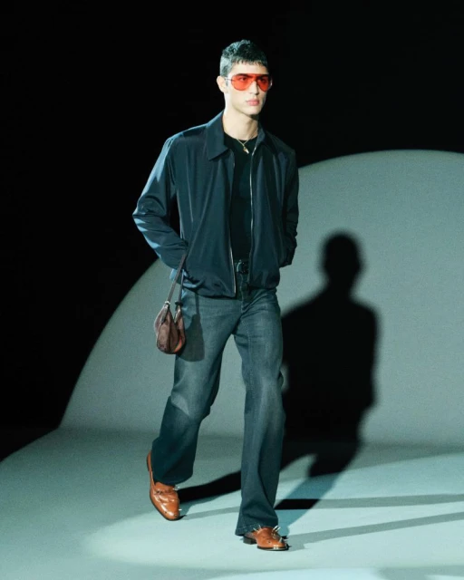 A male model walks on a runway wearing a blue jacket, grey jeans, brown shoes, and red sunglasses in a fashion show.