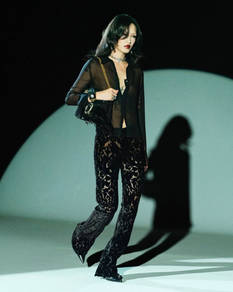 A woman with long dark hair in a sheer black top and patterned pants, holding a black handbag, poses in a studio setting with spotlight.