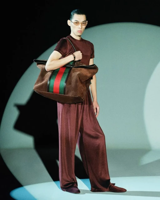 An Asian male model poses in a matching brown top and wide-leg pants, holding a brown Gucci duffel bag with green and red stripes.
