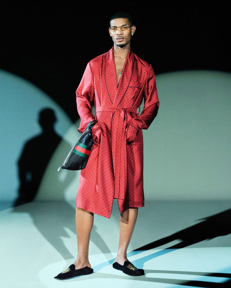 A man models a patterned red robe with black trim, holding a Gucci bag, wearing black velvet loafers and glasses, on a stage with spot lighting.