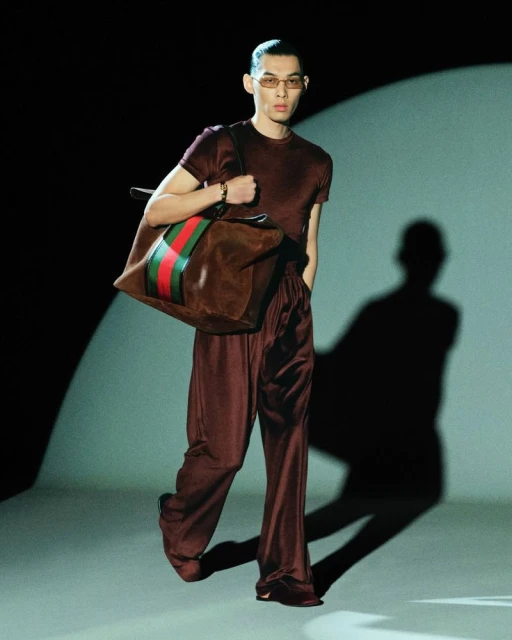 Man with stylish brunette hair wearing a brown satin outfit and sunglasses, carrying a large Gucci duffle bag under studio lighting.