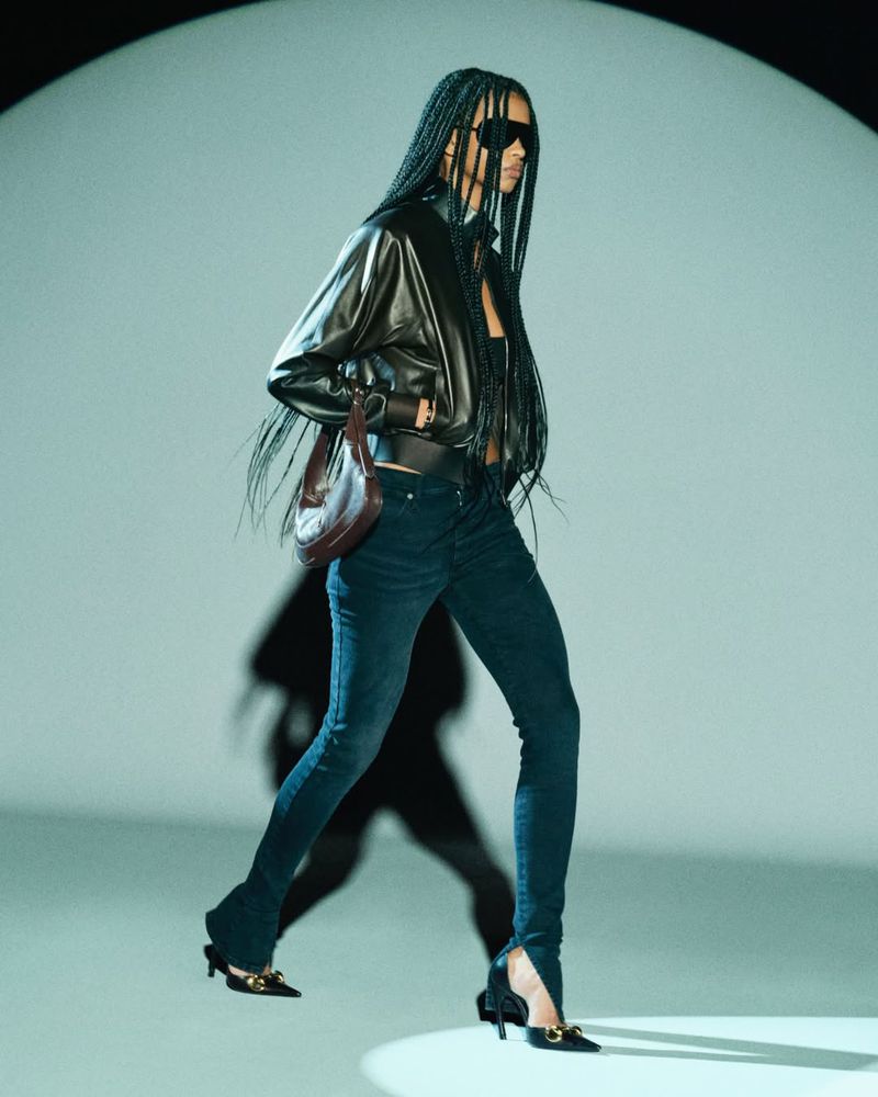 Fashion shot of a woman wearing a leather jacket, jeans, sunglasses, and heels, walking in a lightened studio environment.