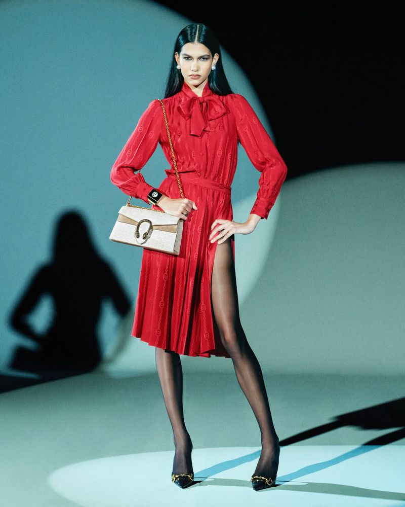 Fashion editorial shot of a tall adult female in a red dress and black tights, carrying a purse in a studio with bright lighting.