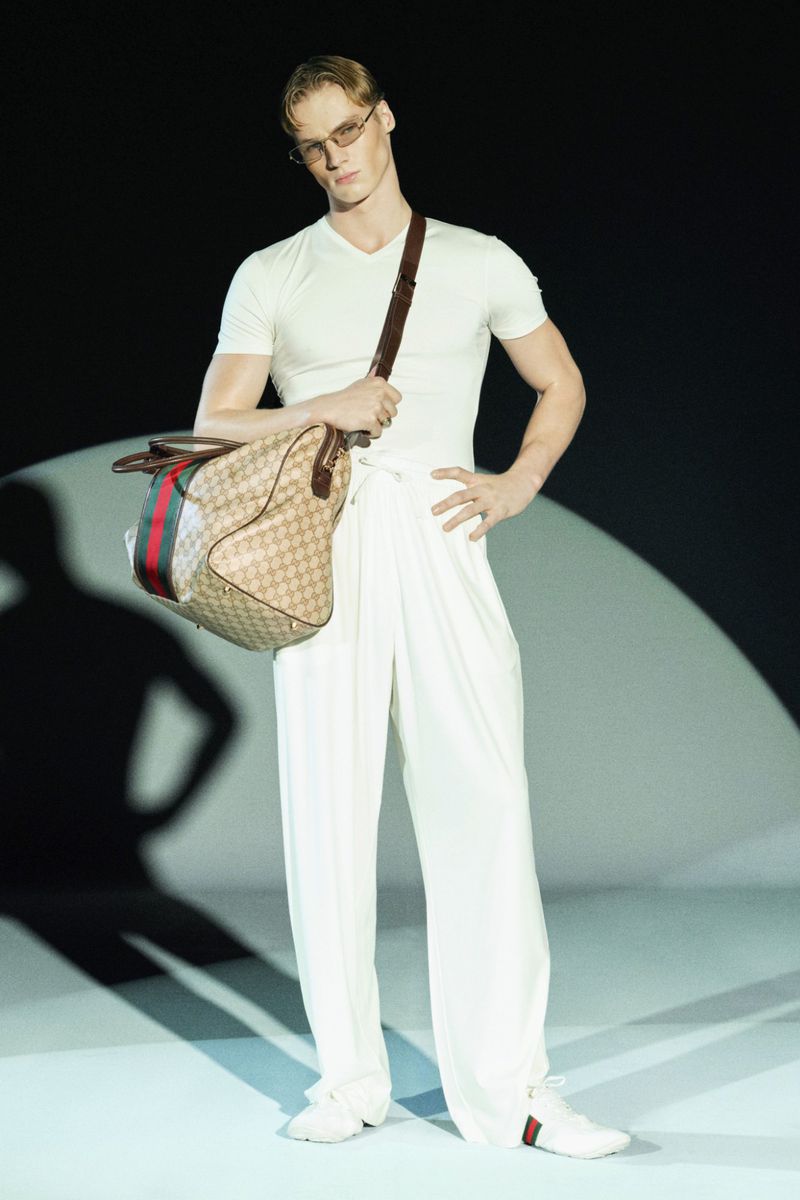 Male fashion model wearing sunglasses and white apparel holds a Gucci duffel bag in a studio setting for a high fashion shoot.