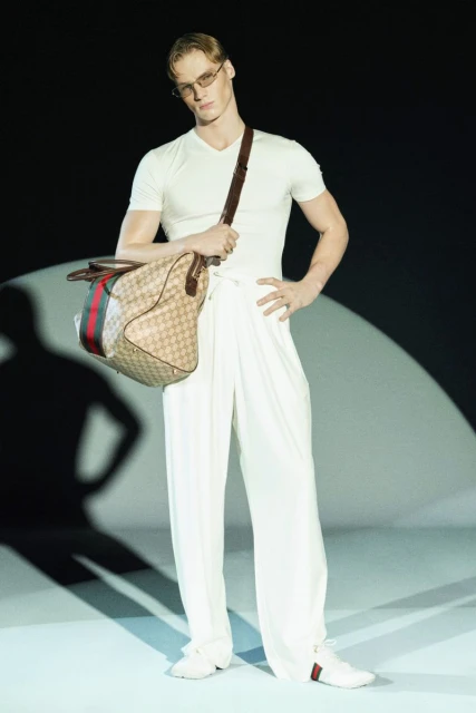 Male fashion model wearing sunglasses and white apparel holds a Gucci duffel bag in a studio setting for a high fashion shoot.