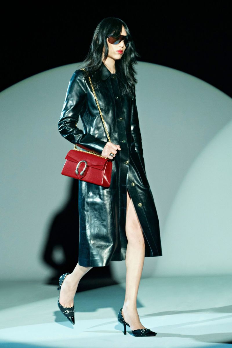 A runway image captures a slender woman in a glossy black trench coat, accessorized with sunglasses and a red purse, walking confidently.