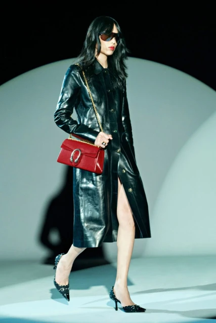 A runway image captures a slender woman in a glossy black trench coat, accessorized with sunglasses and a red purse, walking confidently.