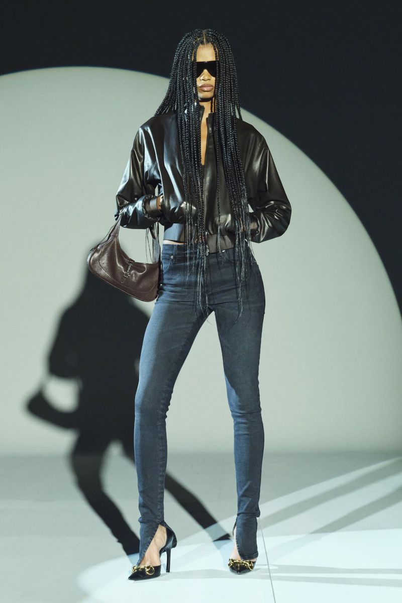 Adult female runway model wearing a black leather jacket and dark denim skinny jeans with a brown purse and black high heels.