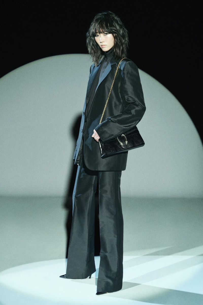 Woman in a black suit with a matching handbag posing against a spotlight, dark background, studio lighting, full-length shot, fashion shoot.