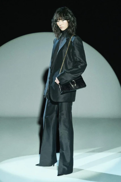Woman in a black suit with a matching handbag posing against a spotlight, dark background, studio lighting, full-length shot, fashion shoot.
